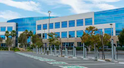 A row of electric vehicle charging stations with dedicated parking spots stands at outdoor parking lot of commercial office building.