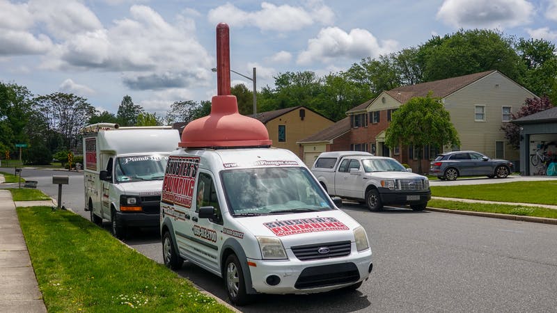 Front of a white plumbing company truck that has a huge plunger on its roof.
